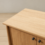 Close-up picture of a light natural teak wood bathroom cabinet with two black round knobs, set against a neutral beige wall and light wood floor.