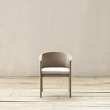 A modern armchair with a beige frame, woven backrest, and a light beige cushioned seat, positioned on a light wood plank floor against a neutral beige wall.