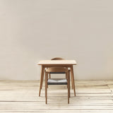 A minimalist dining set consisting of a square dining table and two matching chairs with black woven seats, set against a neutral beige wall and light wood plank floor.