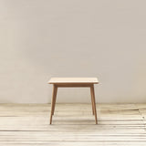 A minimalist light wood square dining table with tapered legs, centered against a neutral beige wall and light wood plank floor