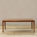 A modern wooden dining table with a warm brown finish, shown on a light wood plank floor against a neutral beige wall.