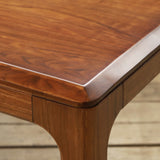 Close-up of a polished wooden table corner with a warm medium-brown wood grain finish, set on a light wood plank floor.
