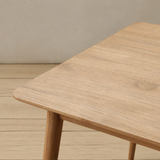 Close-up of a light natural wood table corner with rounded edges and tapered legs, set against a neutral beige wall and wood floor.