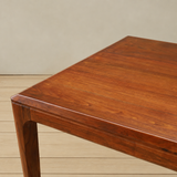 A close-up of a polished reddish-brown wooden table showing the top surface, edge corner, and part of the leg, set against a neutral beige wall and light wood floor.