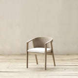 A wooden armchair with a woven backrest and a white cushioned seat, set against a neutral beige wall and light wood plank floor.