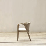 A modern armchair with a light beige frame, woven backrest, and a white cushion, set against a minimalist beige wall and light wood floor.