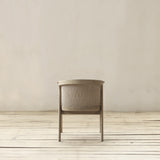A back view of modern chair with a beige wood frame and a woven backrest, set against a neutral beige wall and light wood floor.