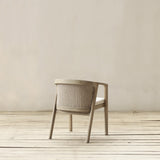 A light beige wooden dining chair with a woven backrest and a beige cushion, set against a neutral beige wall and light wood floor.