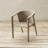 A beige armchair with a woven backrest and slatted seat, positioned on a light wood plank floor against a neutral beige wall.