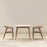 A minimalist dining set featuring a square wooden table and two matching armchairs with light wood frames and black woven seat, set against a neutral beige wall and light wood floor.