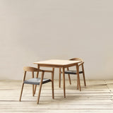 A minimalist dining set featuring a square dining table and two matching chairs with black woven seats, set against a neutral beige wall and light wood floor.