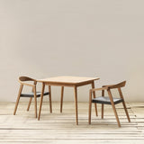 A minimalist dining set featuring a square dining table and two matching armchairs with black woven seats, set against a neutral beige wall and light wood plank floor.