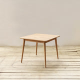 A square wooden dining table with tapered legs, shown in a minimalist room setting with a light beige wall and light wood plank floor.