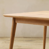 A light natural wood dining table with a square top and tapered legs, shown in a minimalist room setting with a neutral wall and light wood floor.