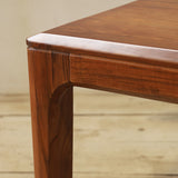 Close-up detail of a wooden dining table corner, showing the polished tabletop, and leg with a warm reddish-brown finish and visible wood grain.
