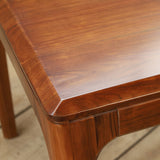 Close-up of a polished reddish-brown wooden table corner showing the grain, beveled edge, and part of the leg on a light wood floor.